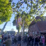 Foto von Personen, die eine geschmückte Birke vor einem Gebäude in traditionellem Ammerländer Baustil aufrichten.